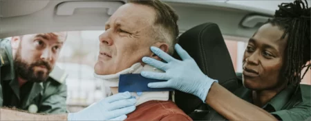 Male seated in car following an accident while medical professionals are placing a brace on his neck.