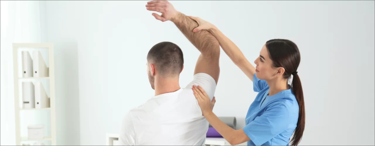 Male patient seated with right arm raised above his head while female physiotherapist assists in therapy on his shoulder.